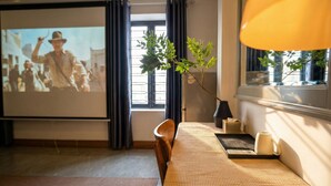 Habitación de hotel con cama, pantalla de proyector mostrando una película, ventana con cortinas, planta y lámpara.