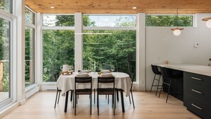 Comedor con mesa dispuesta para cuatro personas, ventanas amplias y zona de cocina con sillas negras y encimera blanca.