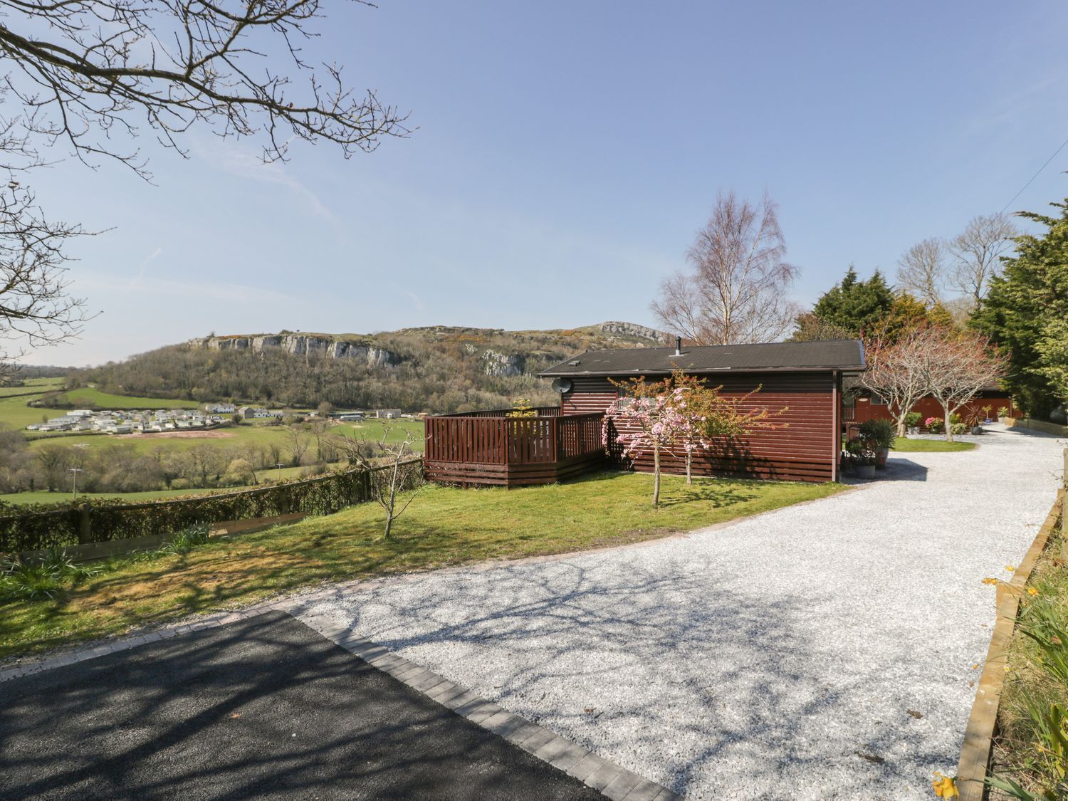 A rural property with a gravel driveway leading to a wooden building, surrounded by greenery and a distant view of a cliff.