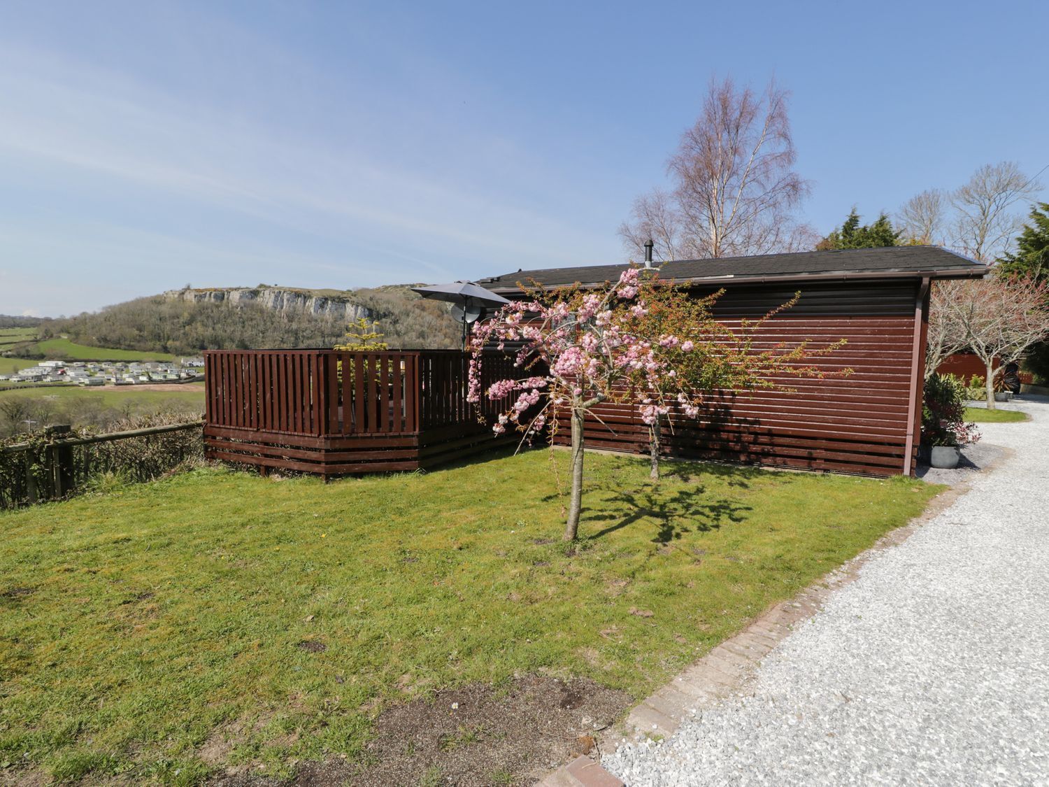 A wooden cabin with a gravel driveway, surrounded by a grassy area and a small tree with pink blossoms.
