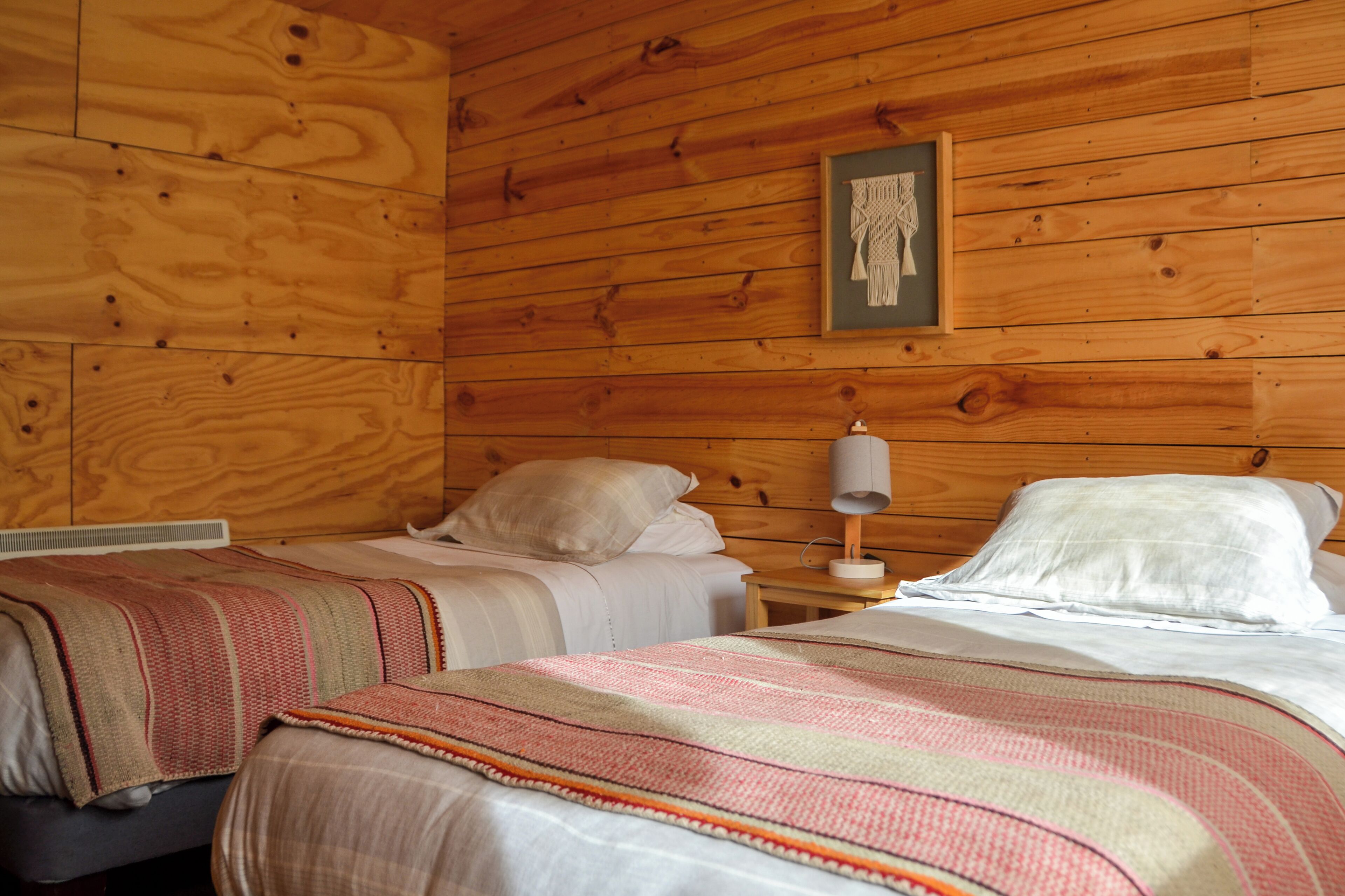 Una habitación con paredes de madera, dos camas con sábanas blancas, una mesita de noche con lámpara y una obra de arte enmarcada en la pared.