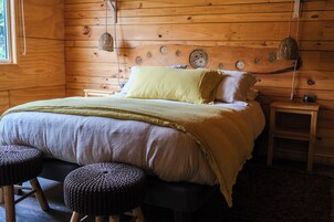 Una habitación de cabaña de madera con una cama, dos taburetes, una mesita de noche y un muro decorado con diseños que evocan el sol.