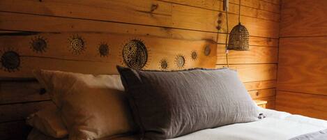 Interior de una cabaña de madera con una cama con dos almohadas y una lámpara de fibra trenzada arriba.