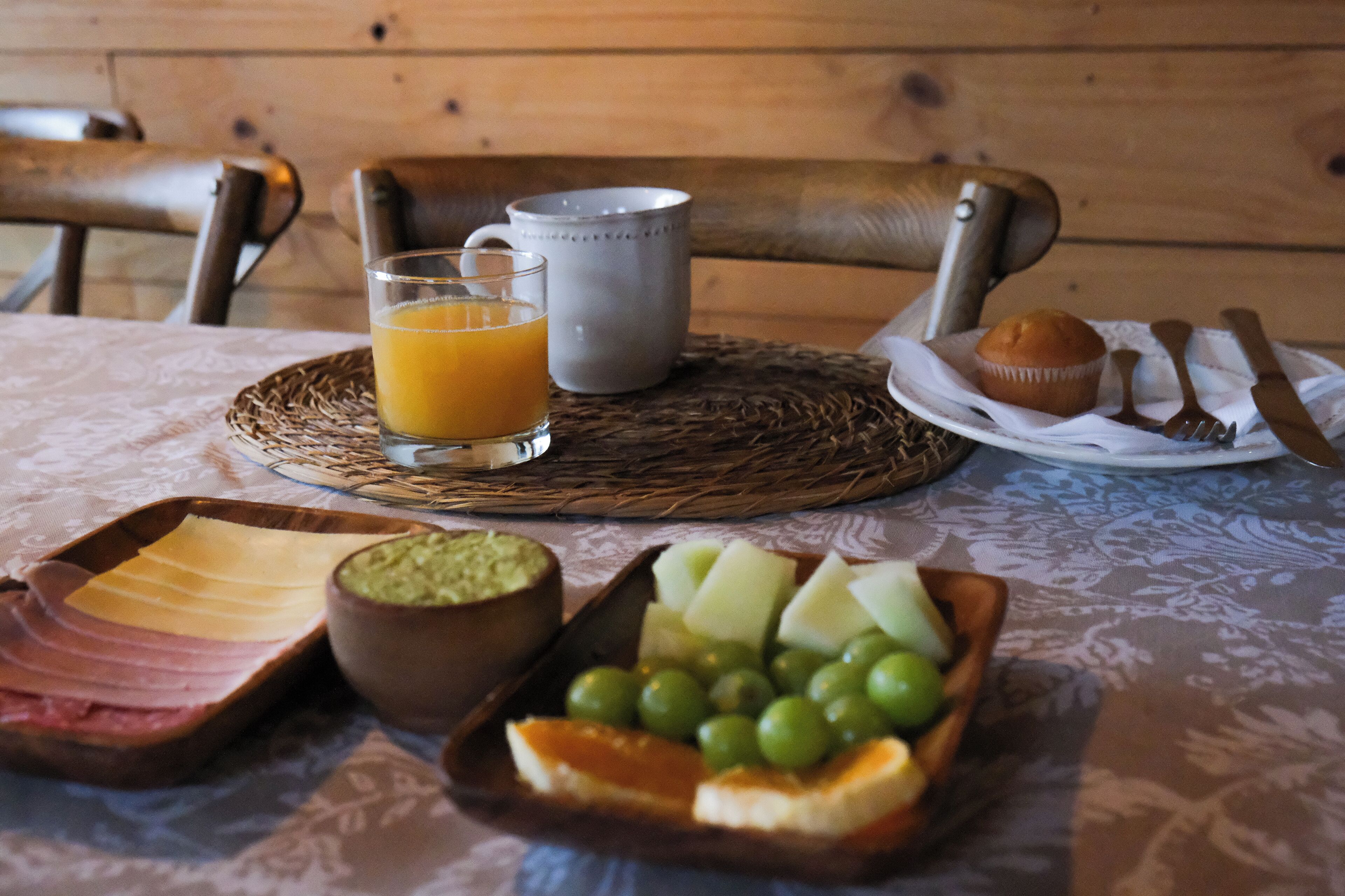 A breakfast setting with a plate of ham, cheese, and fruit, a glass of orange juice, a muffin, and a cup on a woven placemat.