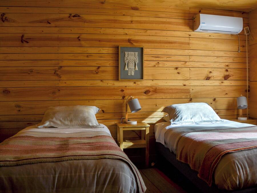 A wooden cabin room with two beds, a framed artwork on the wall, and a ceiling-mounted air conditioner.
