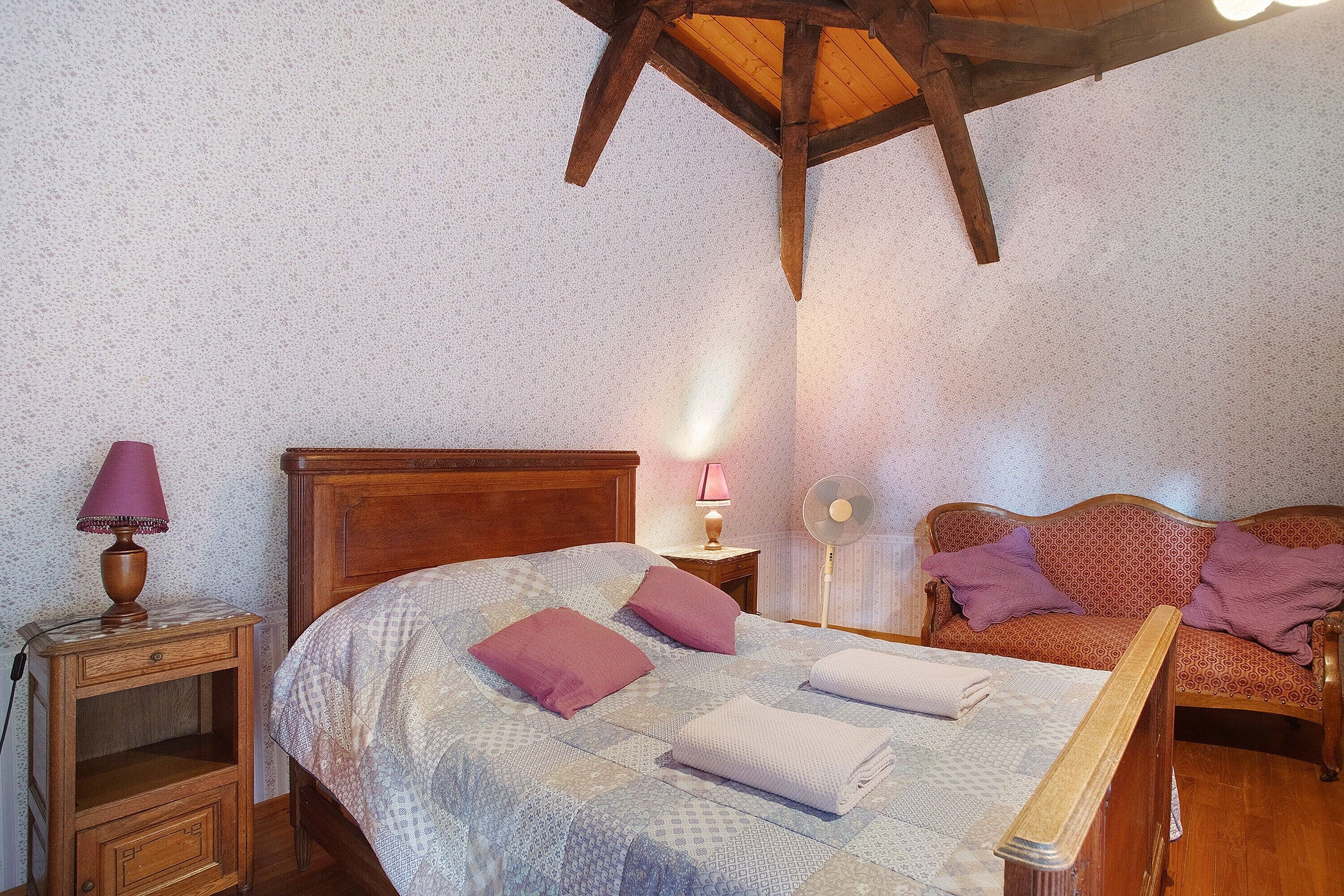 Ein Schlafzimmer mit einem hölzernen Bett, einem Nachttisch, einer Lampe, einem Ventilator und einem gemusterten Sofa.