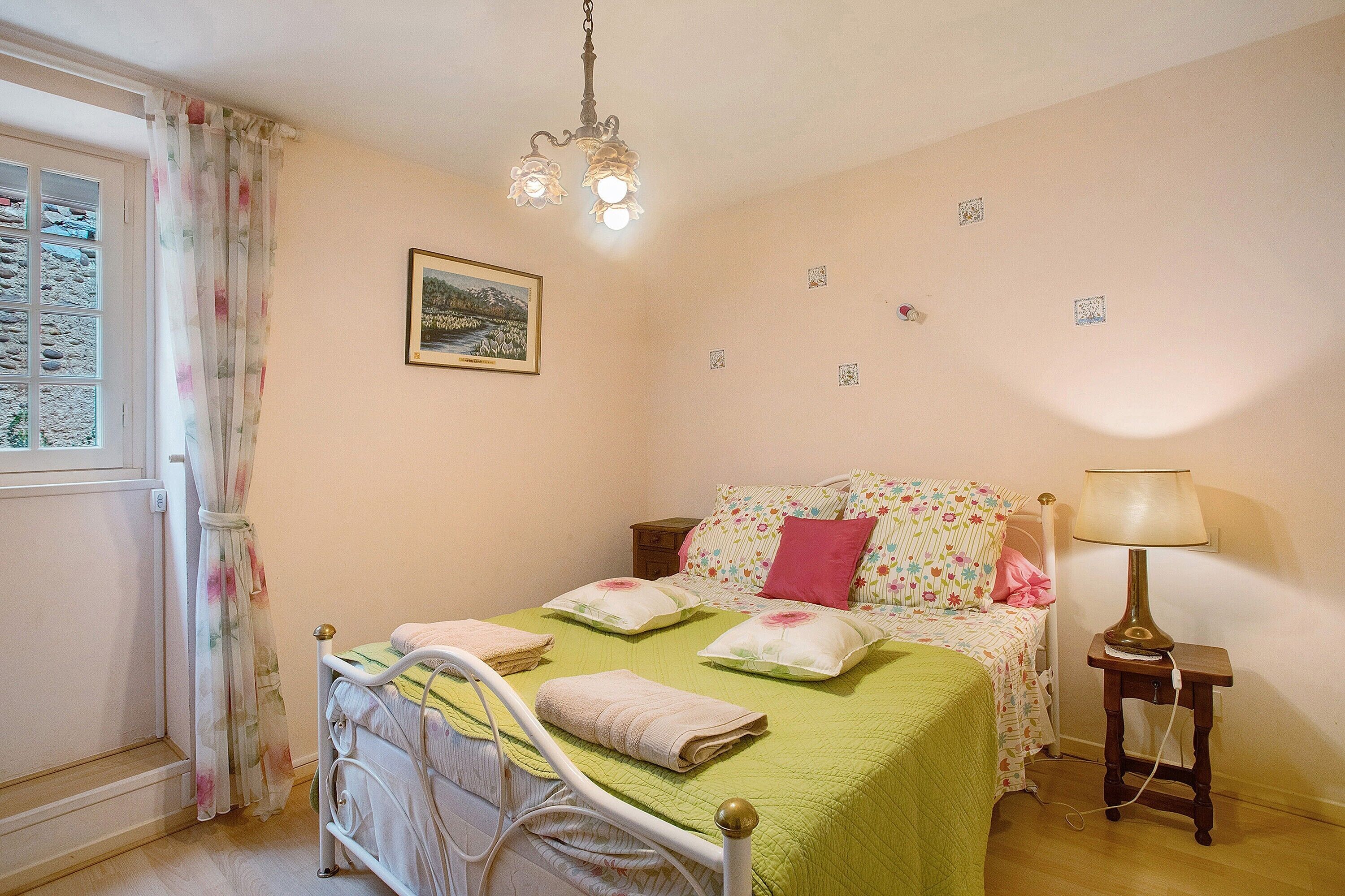 A bedroom with a bed, a bedside table, a lamp, a window with curtains, and a wall-mounted picture.