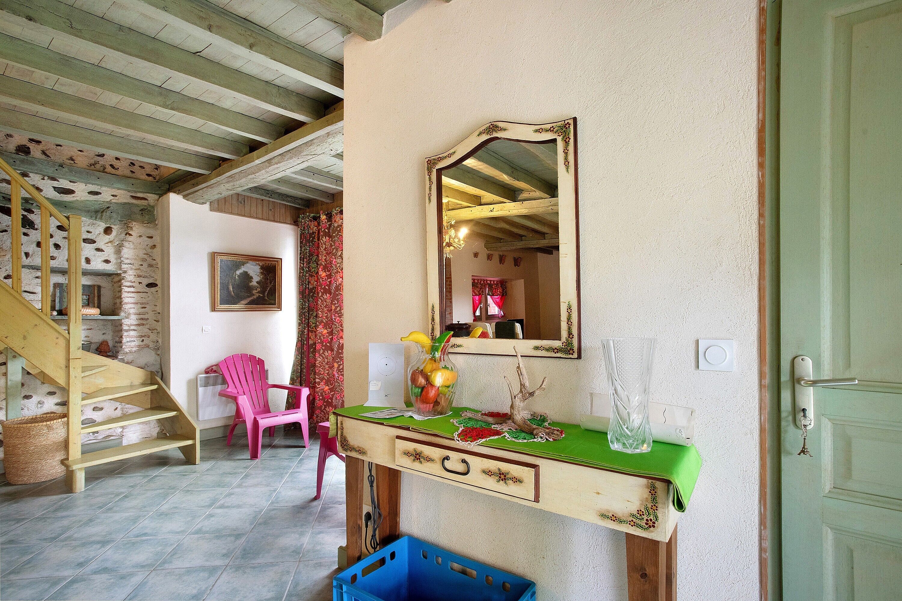 A room with a wooden staircase, a pink chair, a dresser with a mirror, and a vase with fruit.