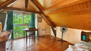 Une pièce avec un plafond en pente, un plancher en bois, une table avec des chaises, un ventilateur et une fenêtre avec des rideaux.