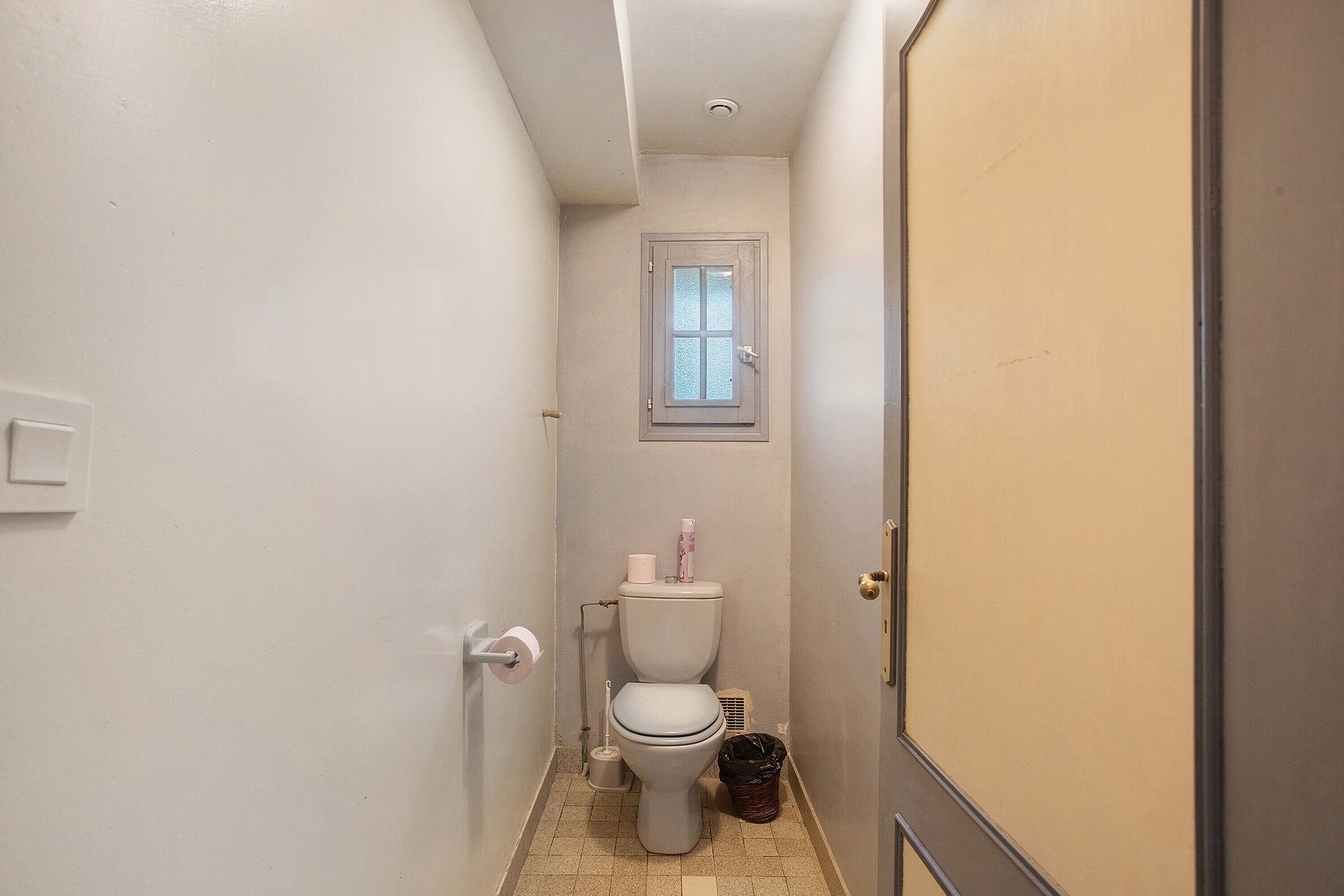 En smal gang, der fører til et badeværelse med toilet, et lille vindue og en dør.