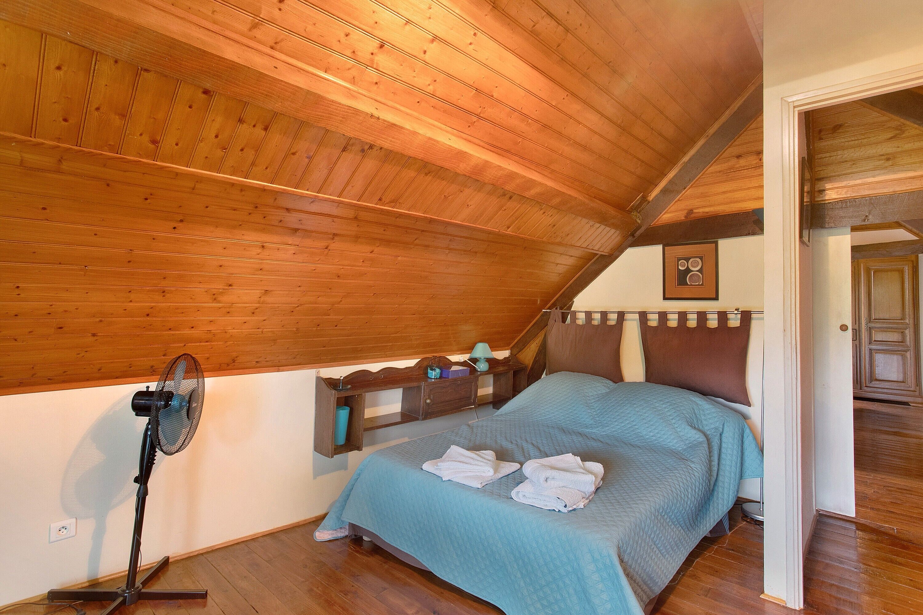 A bedroom with a sloped ceiling, a bed with a blue quilt, a standing fan, and a small shelf with items.