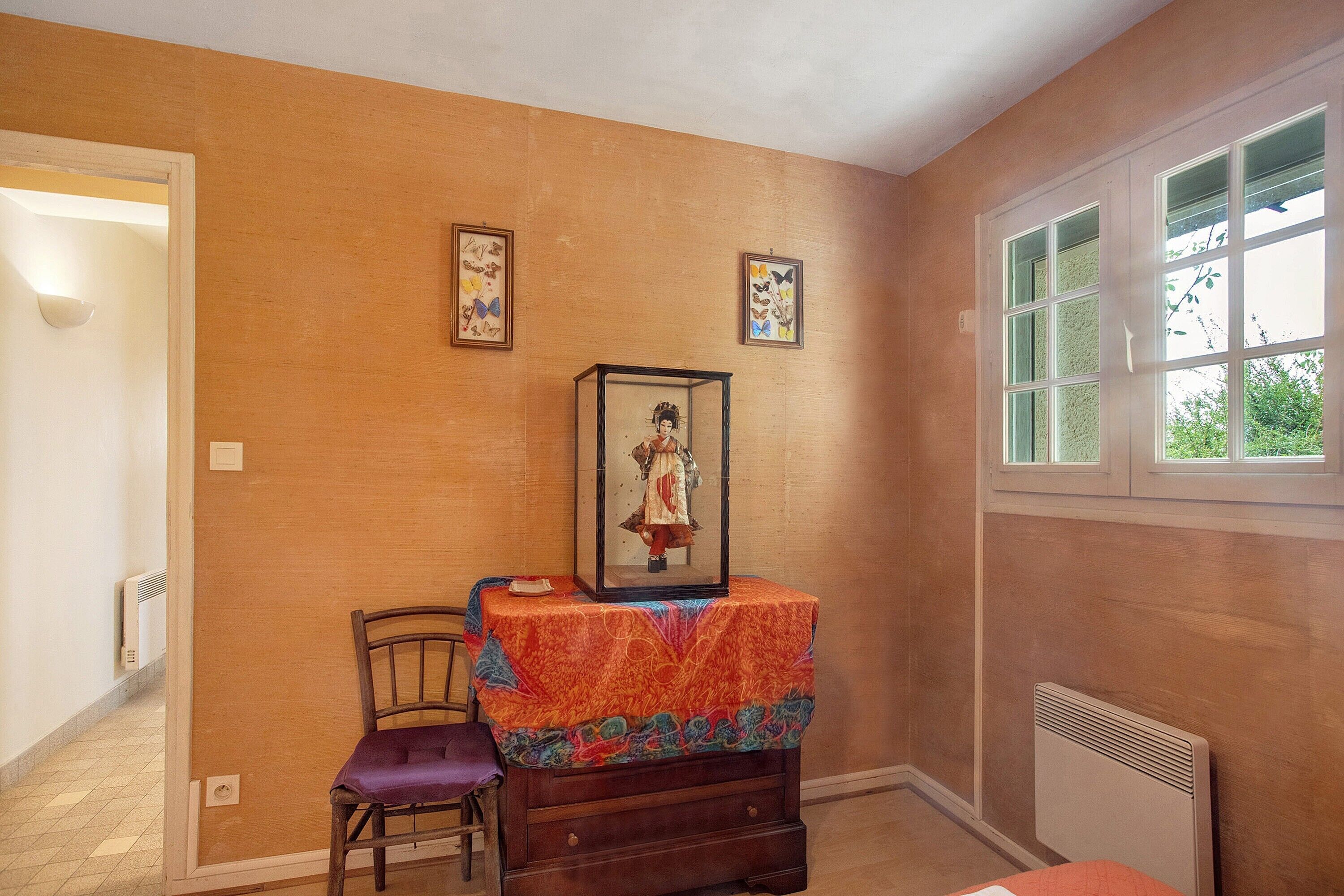 Une pièce avec une chaise en bois, une petite table recouverte d’une nappe colorée et un tableau encadré accroché au mur.