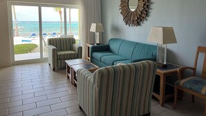 A living room with a view of the ocean, a ceiling fan, a round mirror, and two lamps. - Ocean front 2 bedroom condo - Step onto beach!  private patio w/ amenities (Grand Cayman)