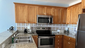 A kitchen with wooden cabinets, a stainless steel microwave, a stove, and a refrigerator. - Ocean front 2 bedroom condo - Step onto beach!  private patio w/ amenities (Grand Cayman)