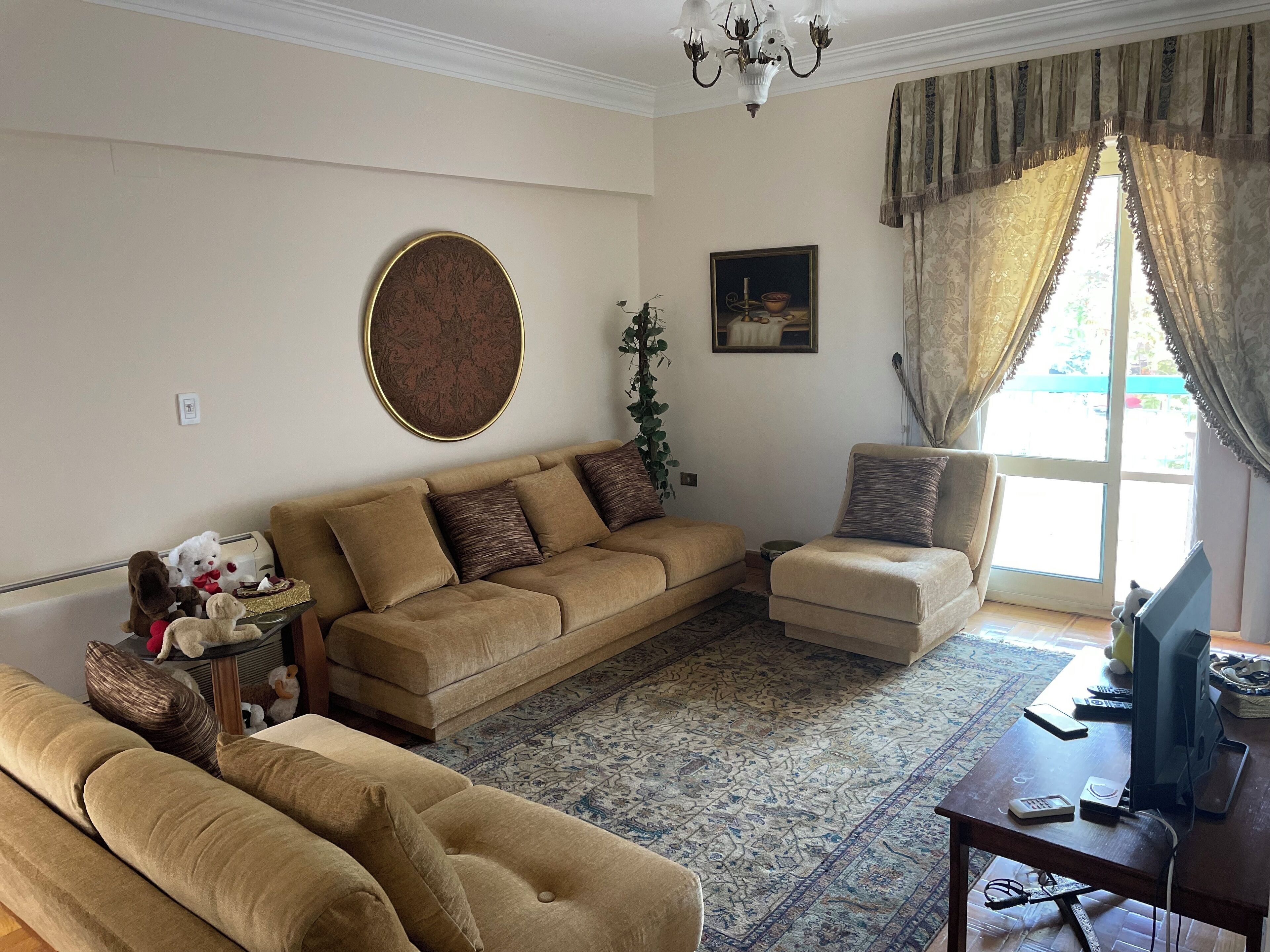A living room with a beige sofa, a matching armchair, a coffee table, a television, and a large window with curtains.