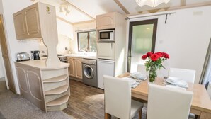 A compact kitchen with a washing machine, a microwave, and a toaster. There is a dining table with chairs and a vase of red flowers. - Springtime Lodge (Abergele)