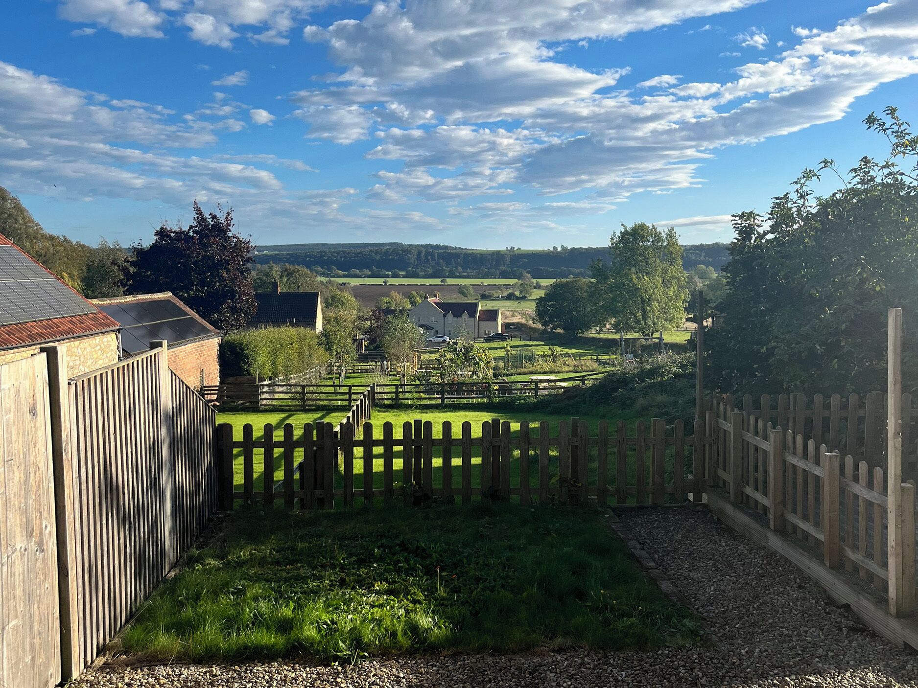 Un paysage rural avec une clôture en bois, un chemin de gravier et des maisons en arrière-plan.