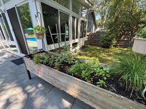 Un jardín elevado de madera con diversas plantas y flores, una valla de madera y una piscina al fondo.