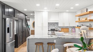 A modern kitchen with white cabinets, a marble island, wooden stools, and stainless steel appliances. - Boardwalk Villas 298 - Community Pool, Lake Front, Great Amenities + Dining! (Miramar Beach)