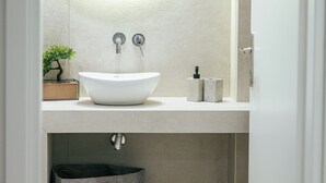 Un baño con un espejo redondo, un lavamanos blanco y un dispensador de toallas. Hay una planta pequeña en la encimera y una bolsa de lavandería con la etiqueta "DHLUX LAUNDRY SERVICE" debajo del lavamanos.