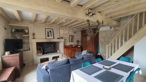 A cozy living room with a fireplace, wooden beams, a sofa, a table with chairs, and a staircase. - La petite massonnière (Beaumont-en-Véron)