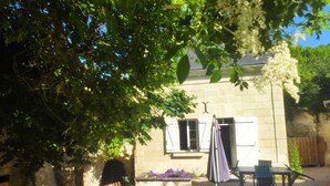 A stone building with a window and a door, surrounded by greenery and a patio area with a table and chairs. - La petite massonnière (Beaumont-en-Véron)