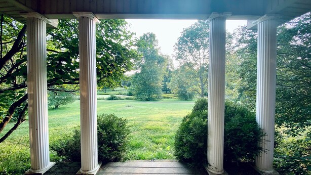 Eine Veranda mit vier weißen Säulen, einem Holzboden und Blick auf ein grasbewachsenes Feld mit Bäumen.