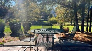 Eine Terrassengruppe im Freien mit rundem Tisch und Stühlen, umgeben von Bäumen und einer Rasenfläche.
