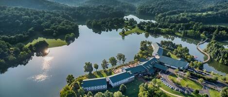 An aerial view of a hotel with a large building, a parking area, and a lake surrounded by forested hills.