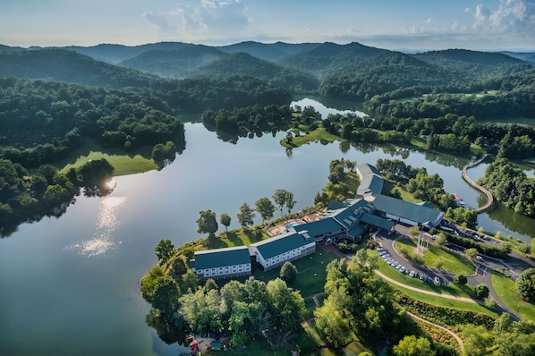 An aerial view of a hotel with a large building, a parking area, and a lake surrounded by forested hills.