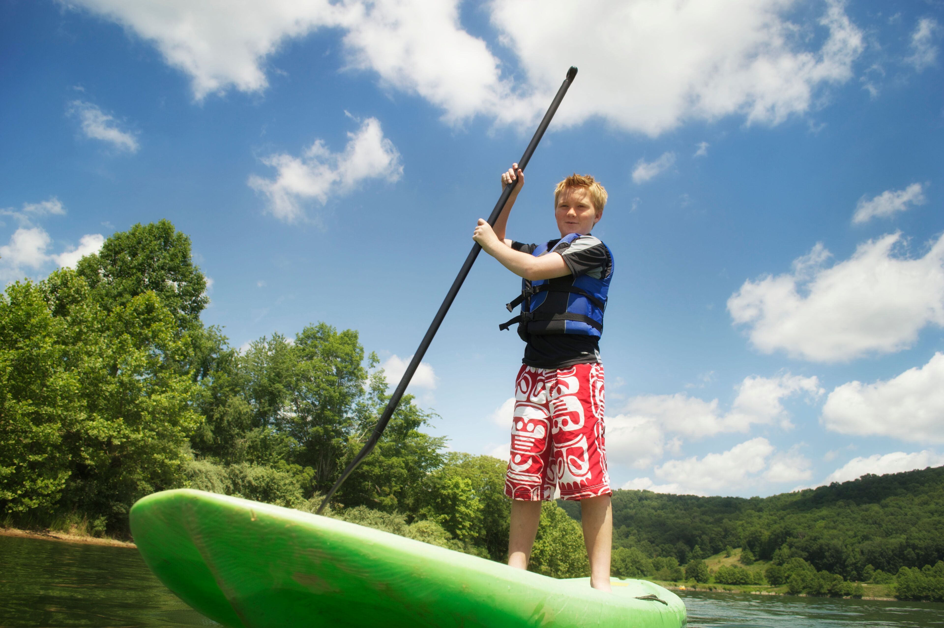 A person standing on a green paddleboard in a body of water with trees and hills in the background.