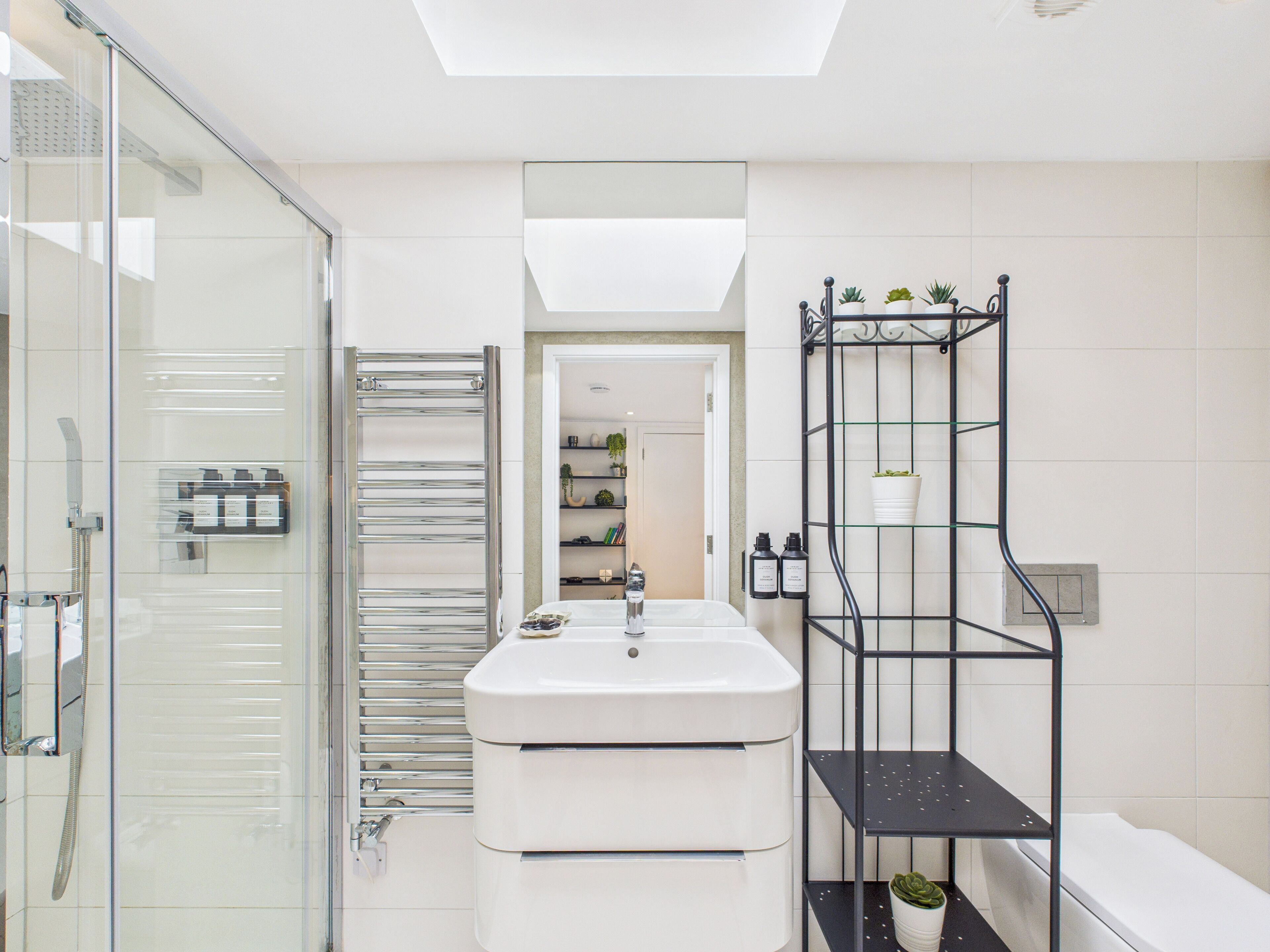 A modern bathroom with a glass shower enclosure, a white sink, and a black shelving unit with plants.