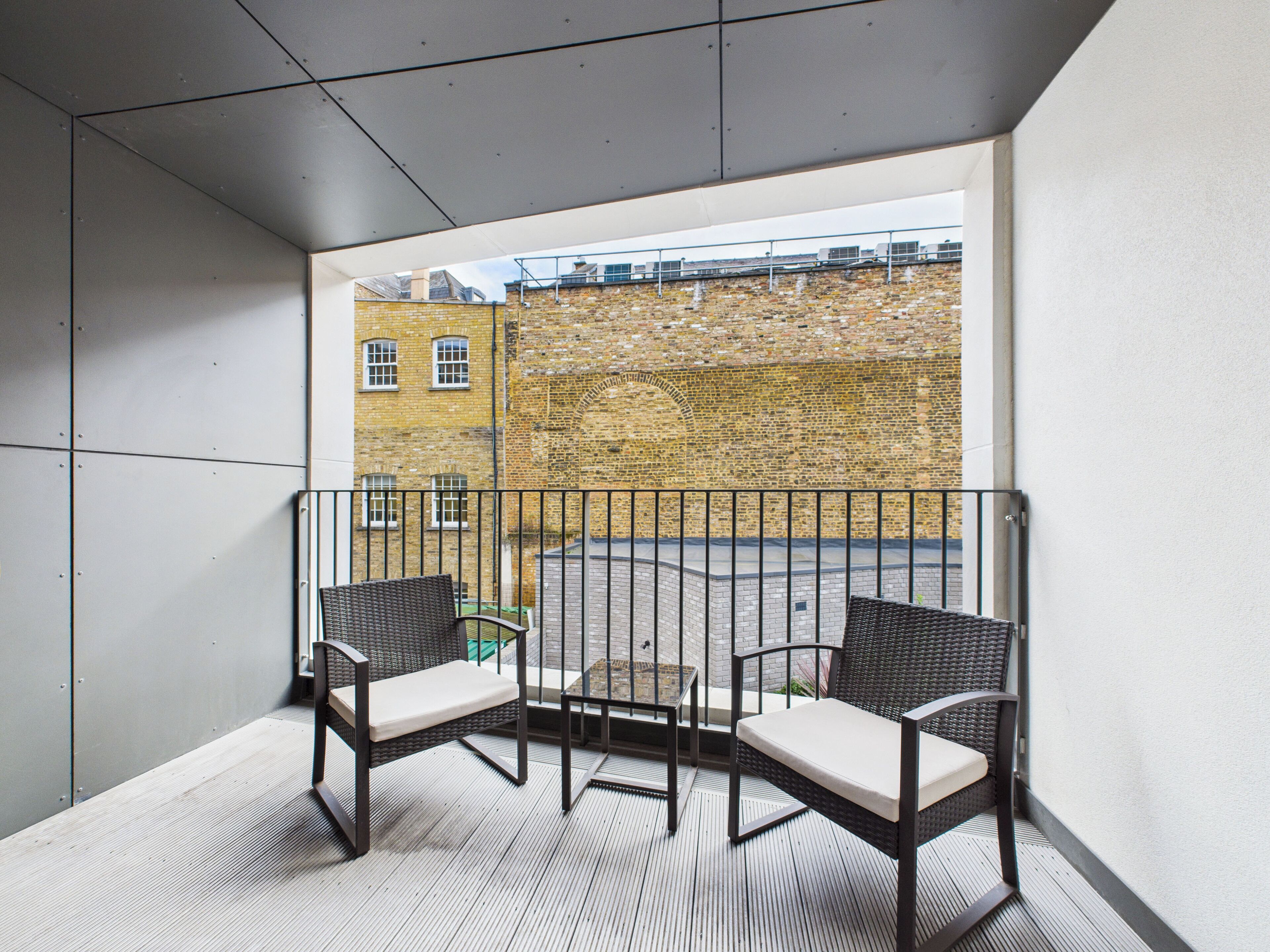 A balcony with two wicker chairs and a small table, overlooking a brick building.