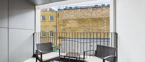 A balcony with two wicker chairs and a small table, overlooking a brick building.