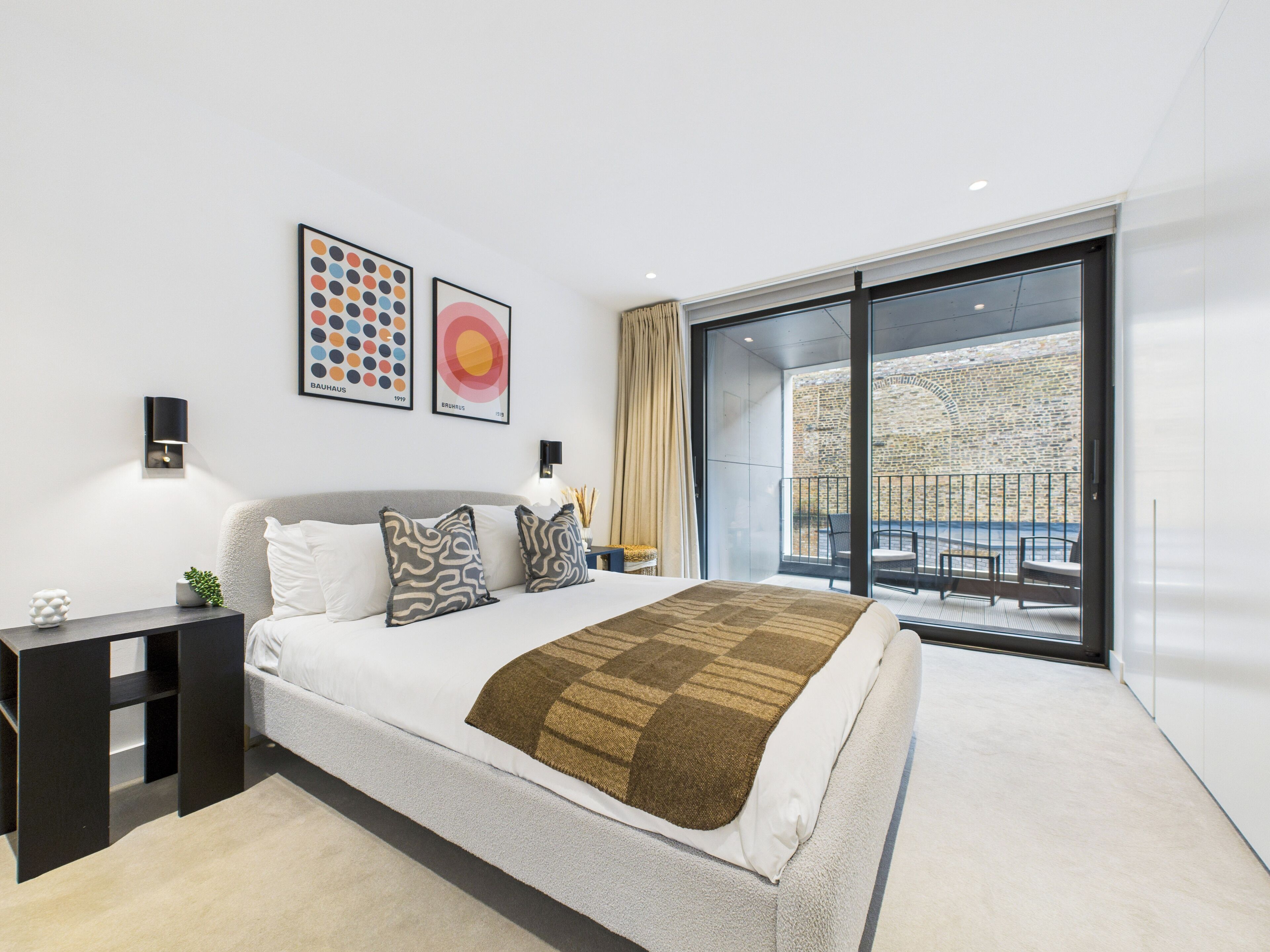 A modern bedroom with a large bed, two wall-mounted lamps, and a view of a stone wall through a sliding glass door.