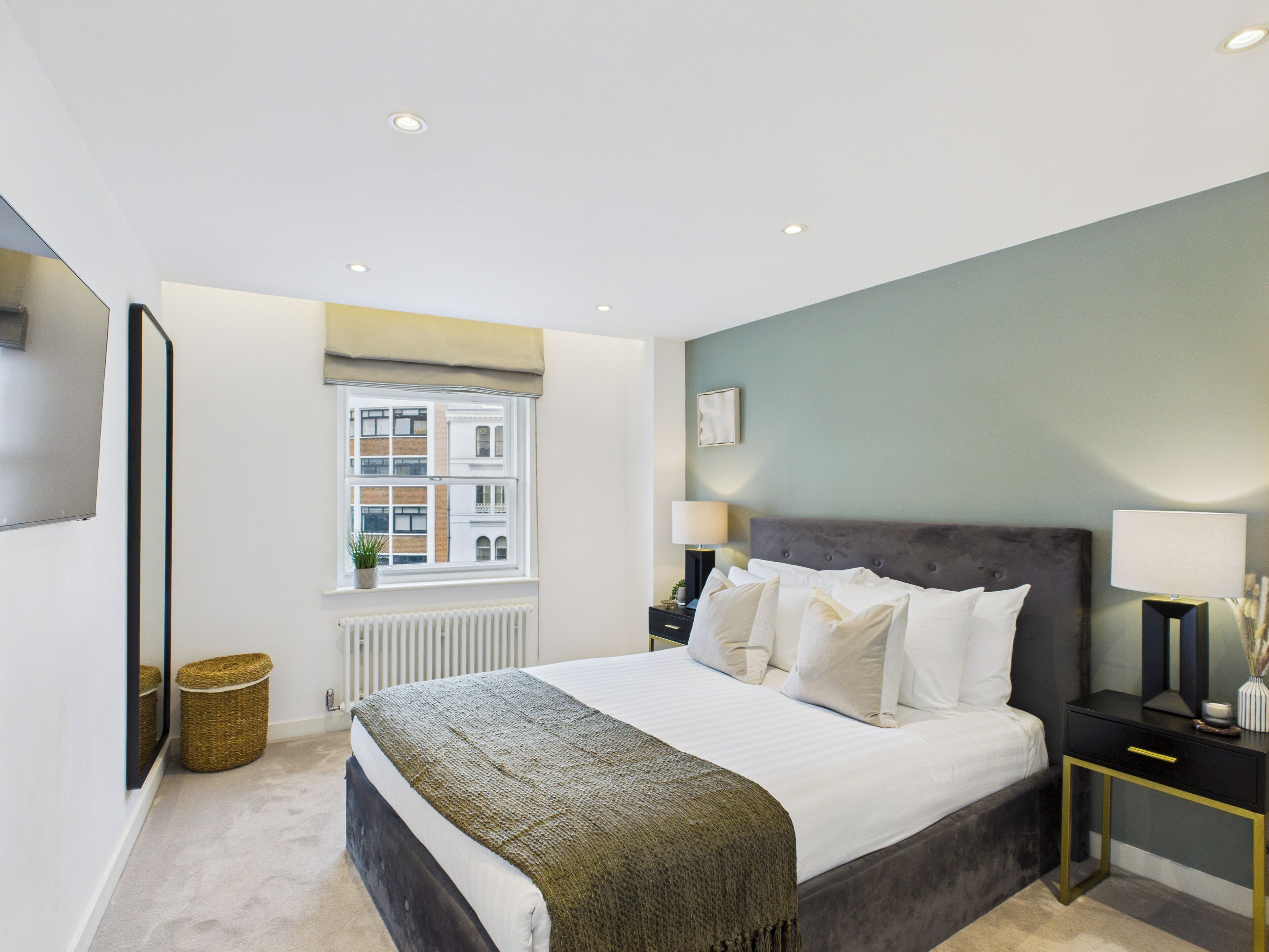 A modern bedroom with a large bed, bedside lamps, a window with a view of buildings, and a radiator.