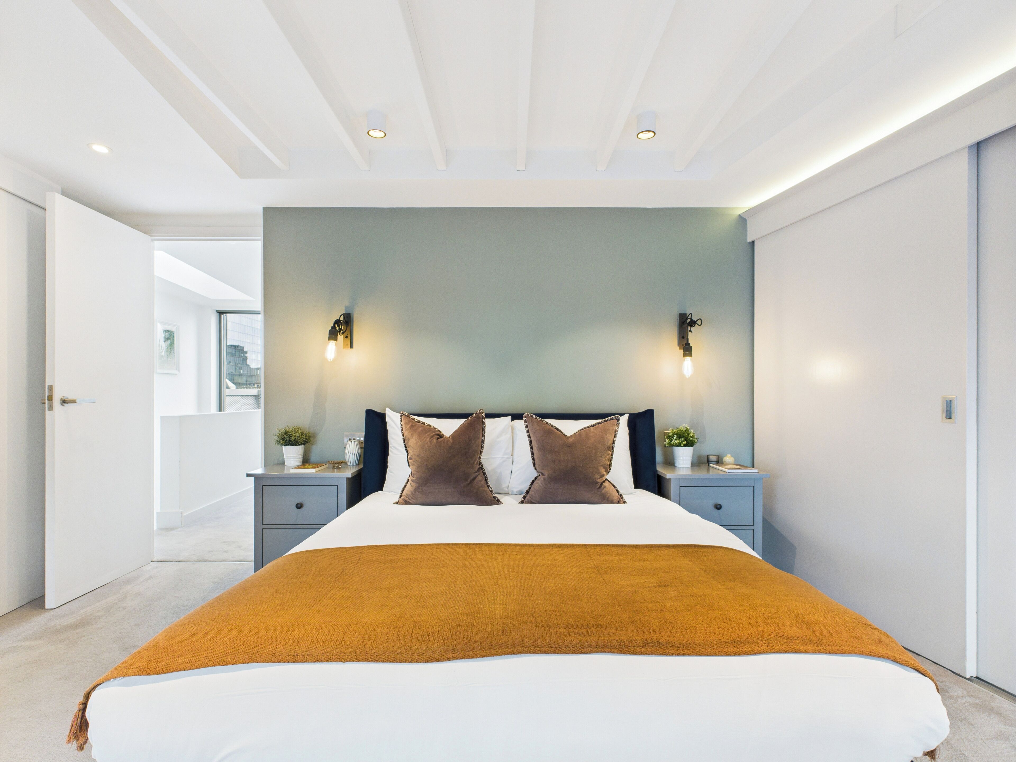 A modern bedroom with a large bed, two bedside tables, a nightstand, a wall-mounted light fixture, and a view of the city through a window.