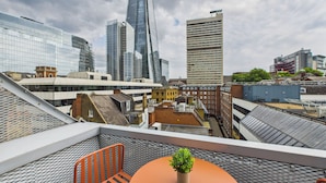 Una terraza en la azotea con una mesa, sillas, una planta en maceta y vistas del horizonte urbano, incluyendo edificios modernos.