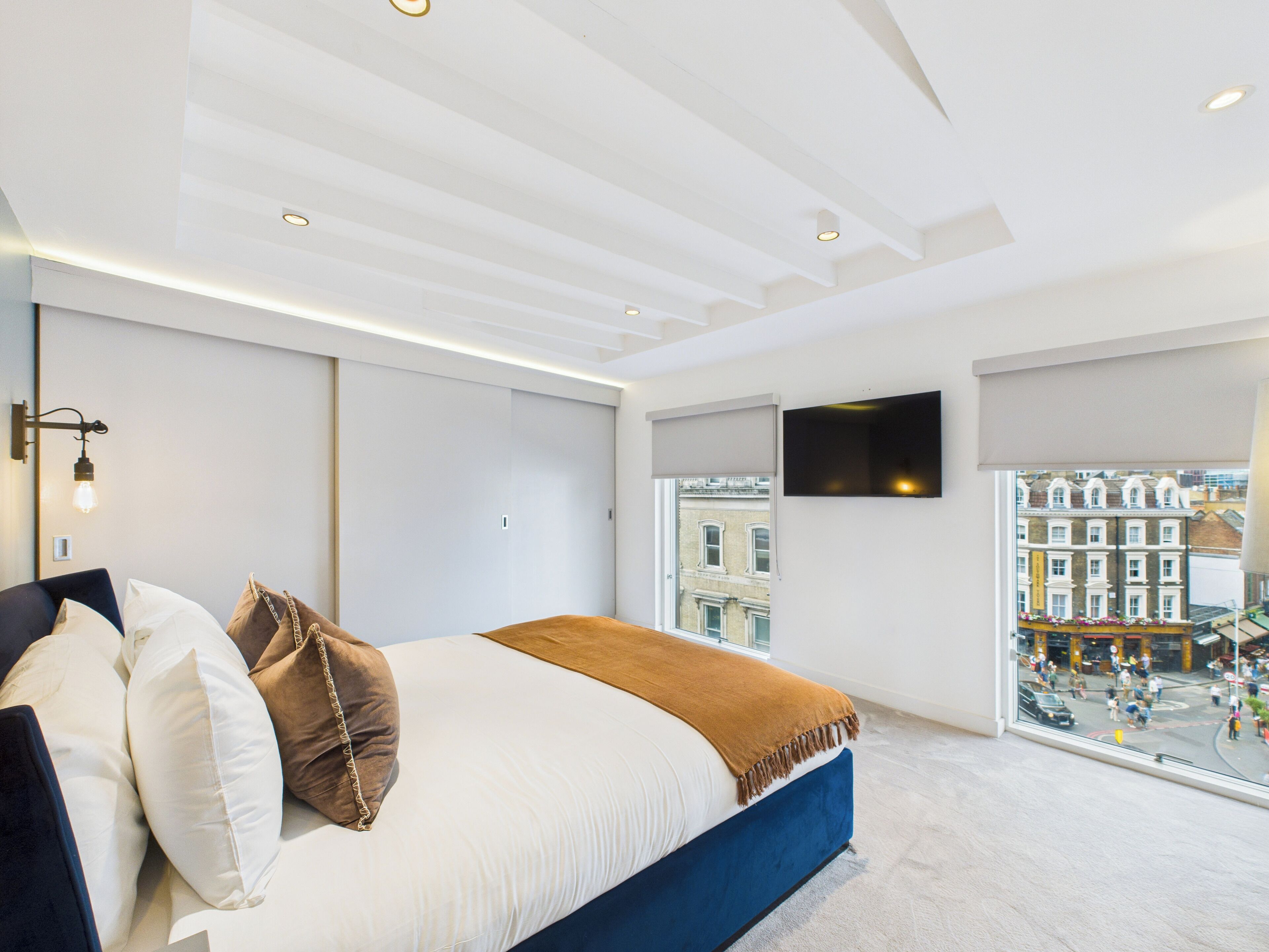 Un dormitorio moderno con una cama grande, un televisor de pantalla plana, una ventana con vistas a la ciudad e iluminación empotrada en el techo.