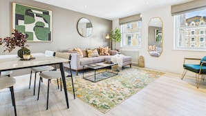 Una sala de estar moderna con sofá, mesa de centro y sillones. Se observa un espejo grande, un jarrón con flores y un cuadro en la pared.