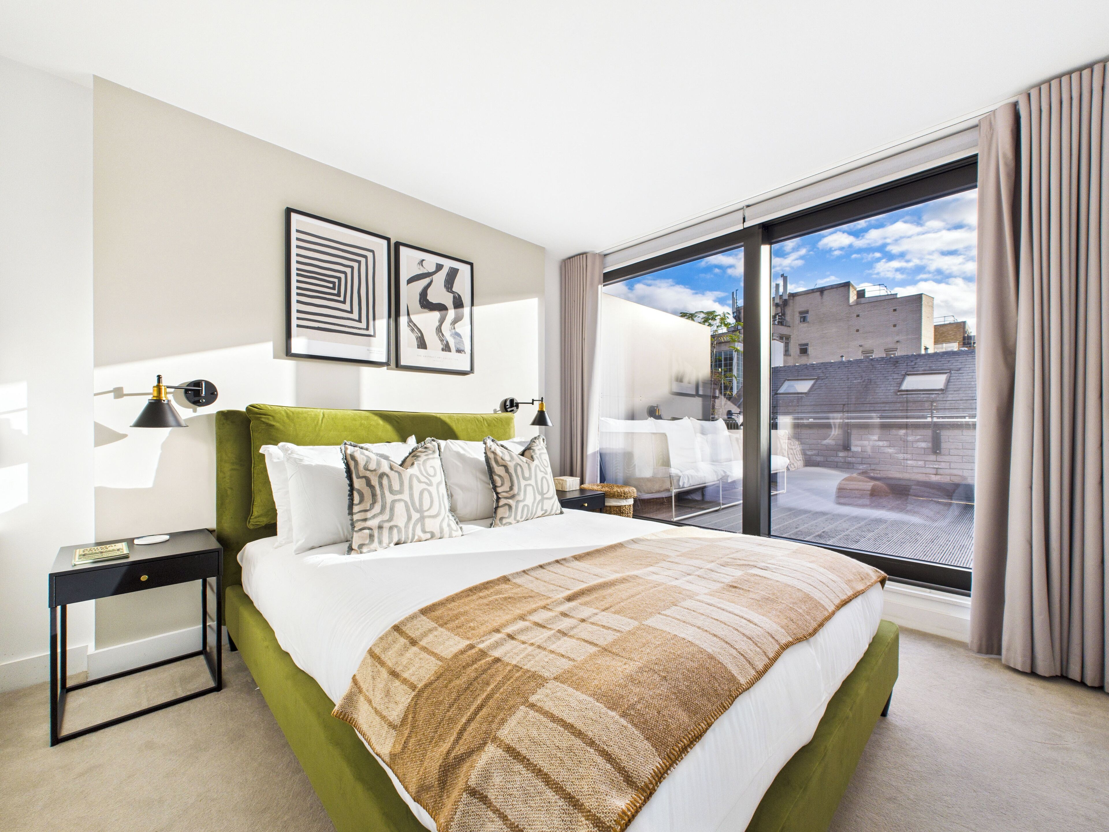 A modern hotel room with a large bed, a green upholstered headboard, and a view of the city through floor-to-ceiling windows.