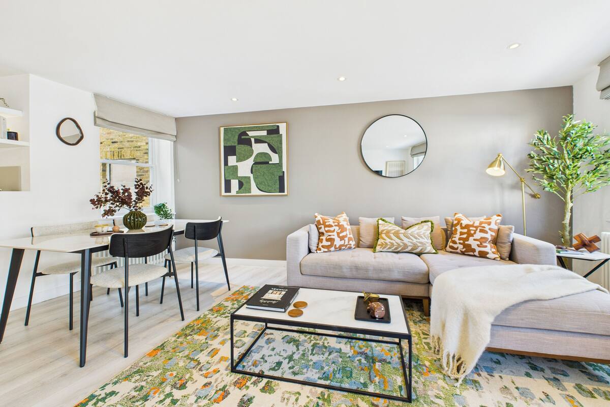Un salon moderne avec un canapé, une table basse et des fauteuils. On y trouve également un coin repas avec une table et des chaises. Un grand tableau abstrait orne le mur, et un miroir est accroché au-dessus du canapé.