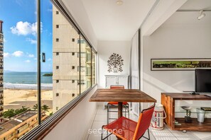 A balcony with a wooden table, red chair, and a beer can. A TV is on a wooden stand, and there's a decorative wall piece. - Beachfront apt BC Center Close to Attract. Aco803 (Balneário Camboriú)