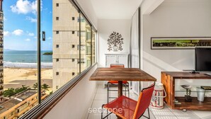 A balcony with a wooden table, red chair, and a beer can. A TV is on a wooden stand, and there's a decorative wall piece. - Beachfront apt BC Center Close to Attract. Aco803 (Balneário Camboriú)