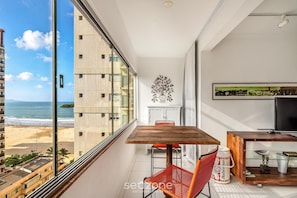 A balcony with a wooden table, red chair, and a beer can. A TV is on a wooden stand, and there's a decorative wall piece. - Beachfront apt BC Center Close to Attract. Aco803 (Balneário Camboriú)