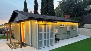Un edificio pequeño de una sola planta con techo inclinado, un porche cubierto con luces colgantes y una puerta que da a un espacio interior.