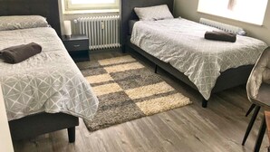 Quarto pequeno e moderno com duas camas, piso de madeira e tapete quadriculado.