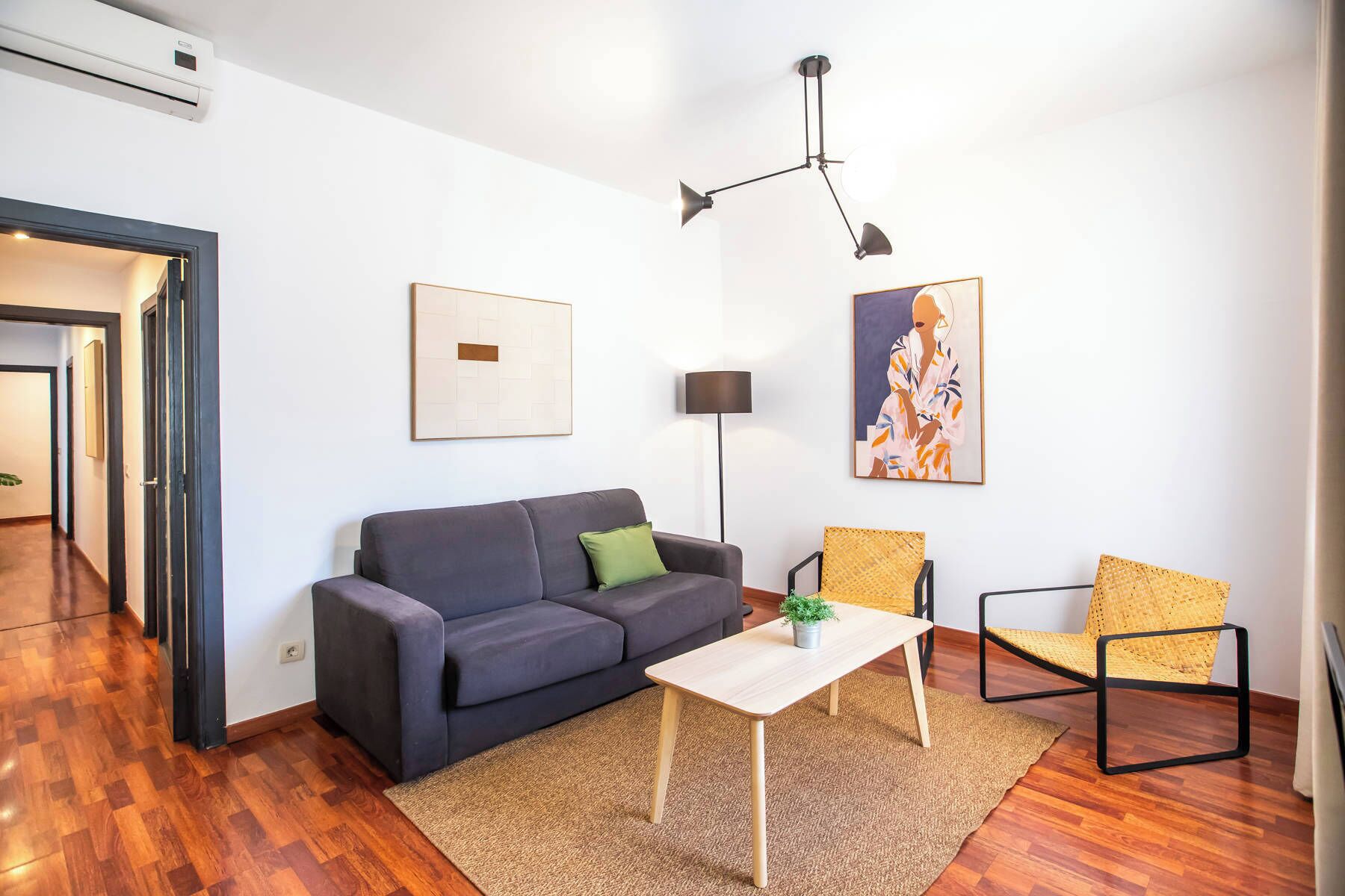 A modern living room with a grey sofa, a small wooden coffee table, two wicker chairs, a floor lamp, and artwork on the walls.