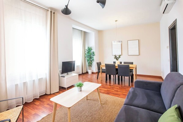 A modern living room with a sofa, a TV, a dining table with chairs, and a potted plant.