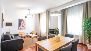 Ein modernes Wohnzimmer mit einer Couch, zwei Sesseln, einem Couchtisch und einem Esstisch. Ein Fernseher, eine Stehlampe und ein Gemälde sind an der Wand zu sehen.